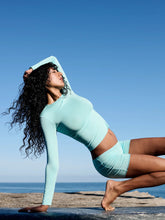 Load image into Gallery viewer, Woman in light blue bamboo  long sleeve athletic wear performing a yoga pose on a rocky beach with clear blue sky.